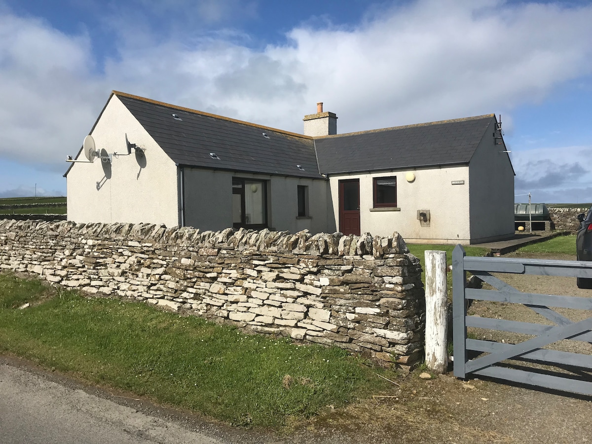 The cottage stands surrounded by a low stone wall and a lush green lawn, with a gravel pathway leading to the entrance. A few satellite dishes are mounted on the roof, and the clear blue sky adds to the serene setting.