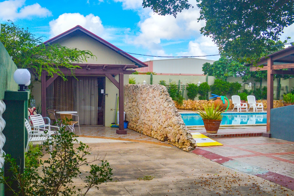A private pool area is surrounded by a stone wall, with a set of six lounge chairs positioned by the water. A small patio space includes a dining table and is framed by lush greenery. The entrance to the accommodation is visible, leading into the living space.