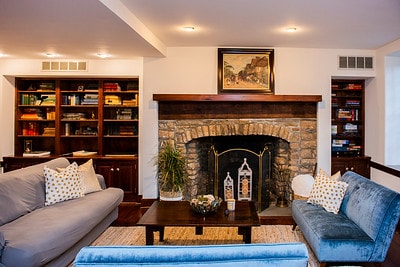 A warm living area features a stone fireplace with a wooden mantle, flanked by built-in bookshelves filled with books. Comfortable seating, including a grey sofa and a blue armchair, is arranged around a central coffee table, enhancing the inviting ambiance.