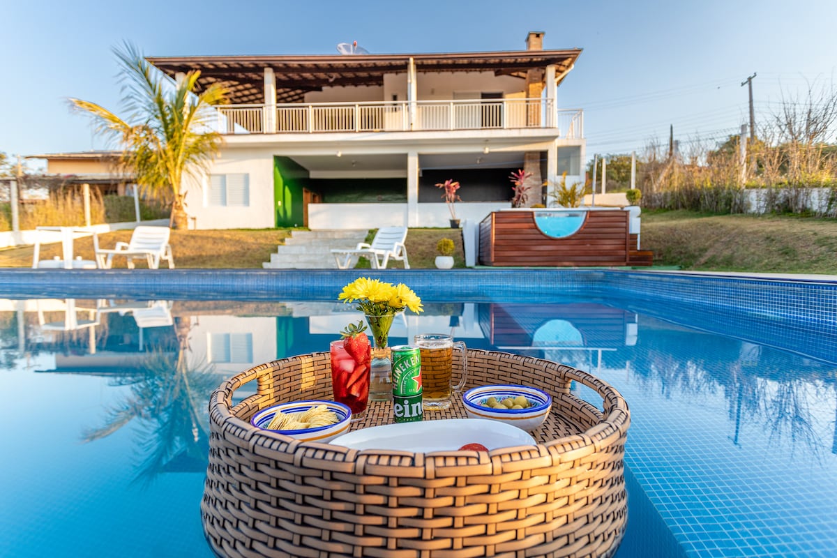 A spacious outdoor pool features a wooden tray holding small dishes and beverages beside vibrant yellow flowers. In the background, a two-story house is visible, showcasing a large balcony and greenery. Sun loungers are arranged near the water's edge, offering a tranquil setting.