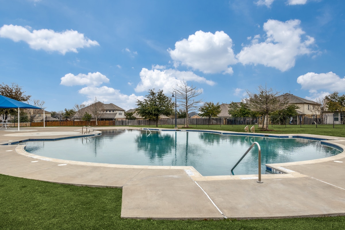 A community pool is featured, surrounded by well-maintained grassy areas. The pool's shallow entry is visible, alongside steps leading into the water. Modern homes are seen in the background against a bright, blue sky dotted with fluffy clouds.