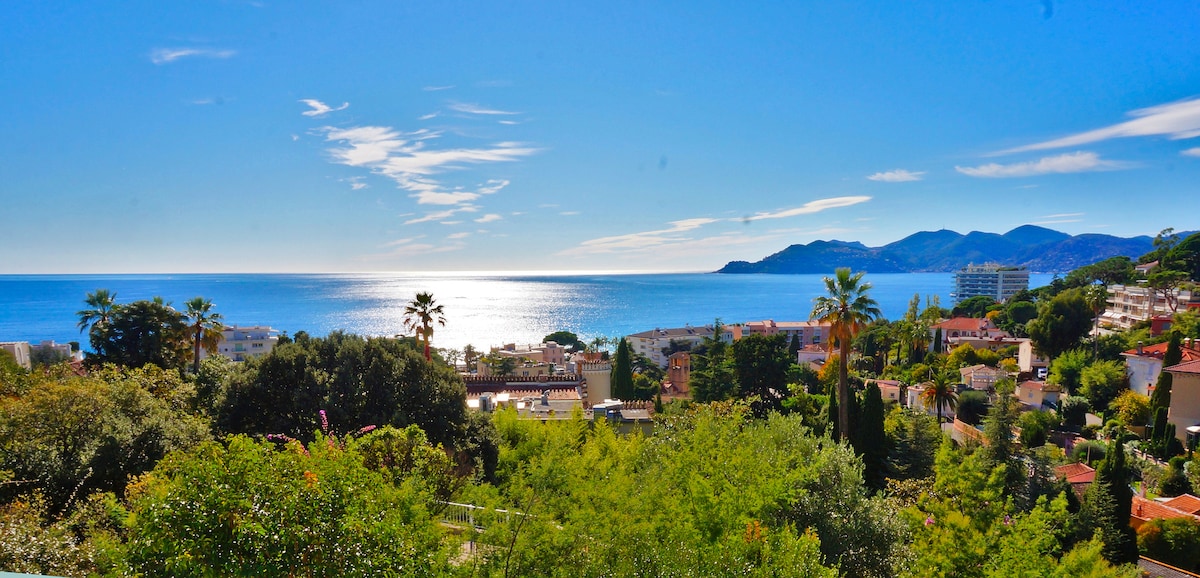 A panoramic view of the Mediterranean Sea is displayed, framed by lush greenery and residential buildings below. The sky is clear and bright, with gentle clouds reflecting sunlight. The distant hills and coastline are visible, adding to the serene coastal landscape.