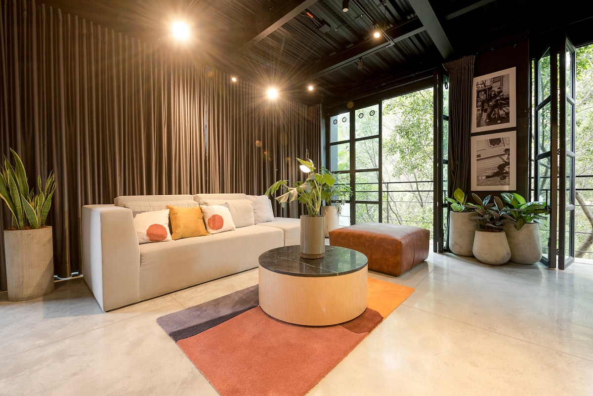 A modern living area features a light-colored sofa with decorative pillows, complemented by a round coffee table. Large windows offer views of lush greenery, while stylish plants adorn the space. Smooth concrete flooring and textured curtains enhance the contemporary aesthetic.