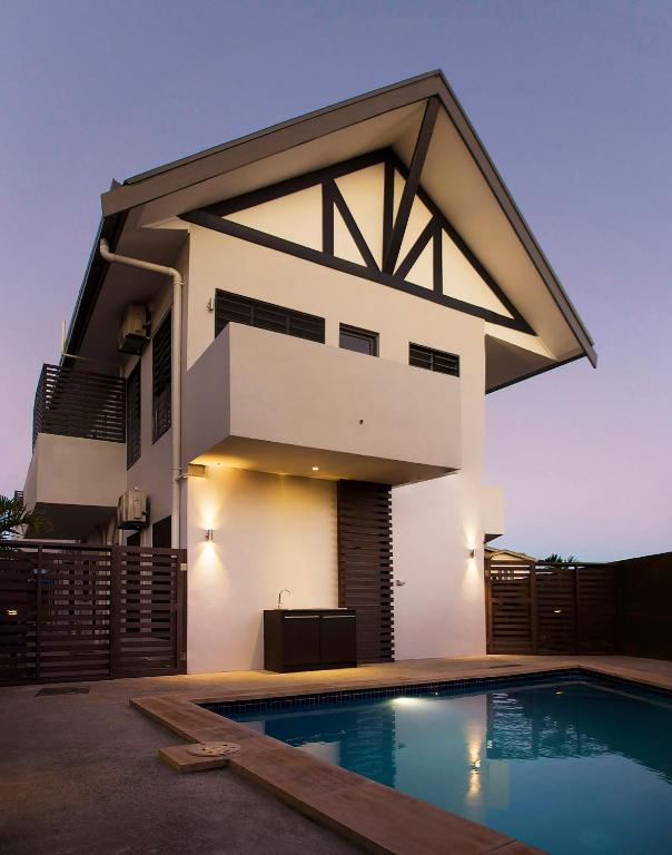 An elegant two-story building is depicted with a distinct peaked roof and modern design. A private swimming pool is visible in the foreground, bordered by a wooden deck. The exterior is illuminated softly, highlighting the structure's features against a twilight backdrop.