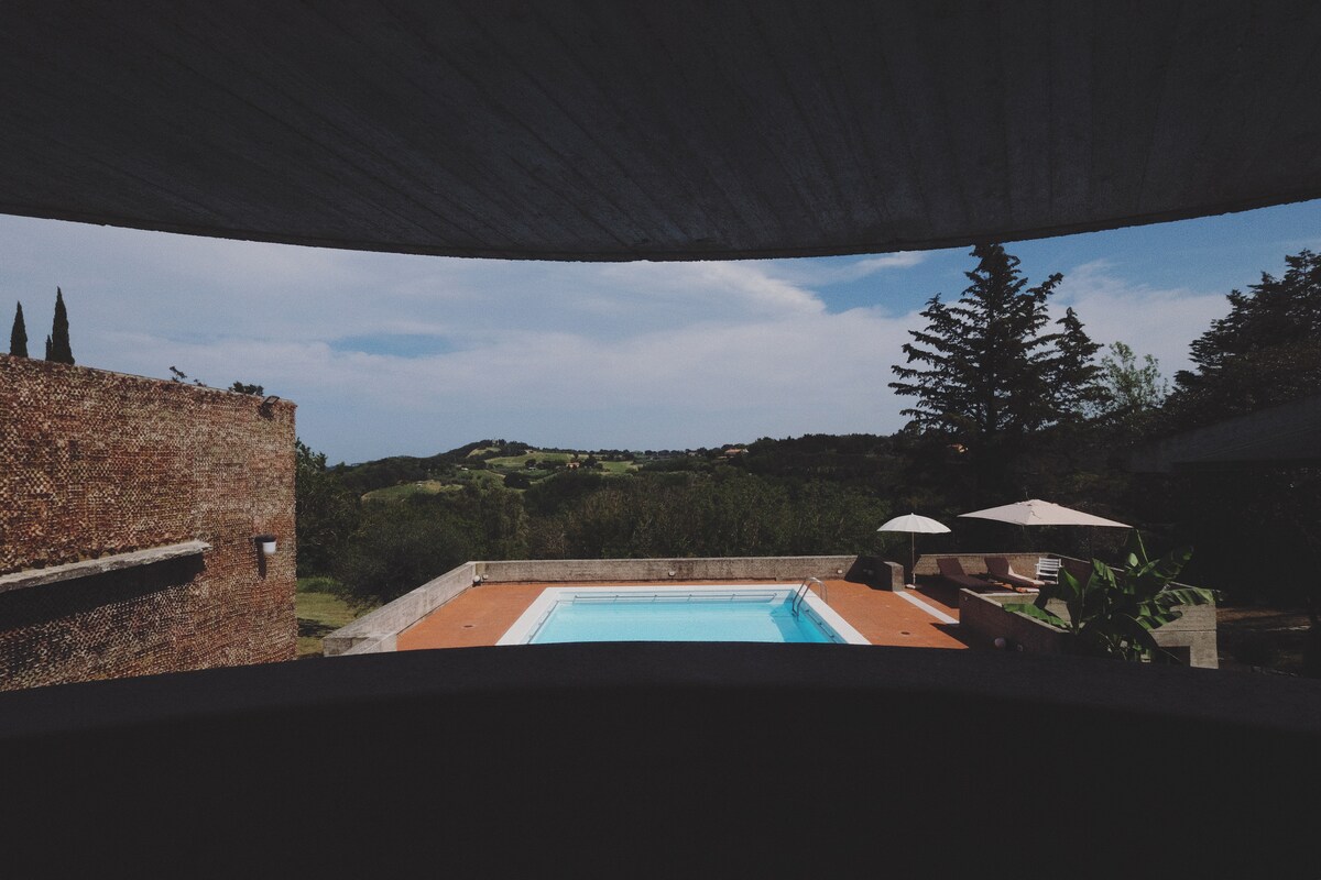 A scenic view from an architectural overhang reveals a refreshing swimming pool surrounded by a sun deck. Lush greenery and rolling hills are seen beyond the pool, enhancing the tranquil outdoor setting. Umbrellas provide shade, creating a calm retreat for relaxation.