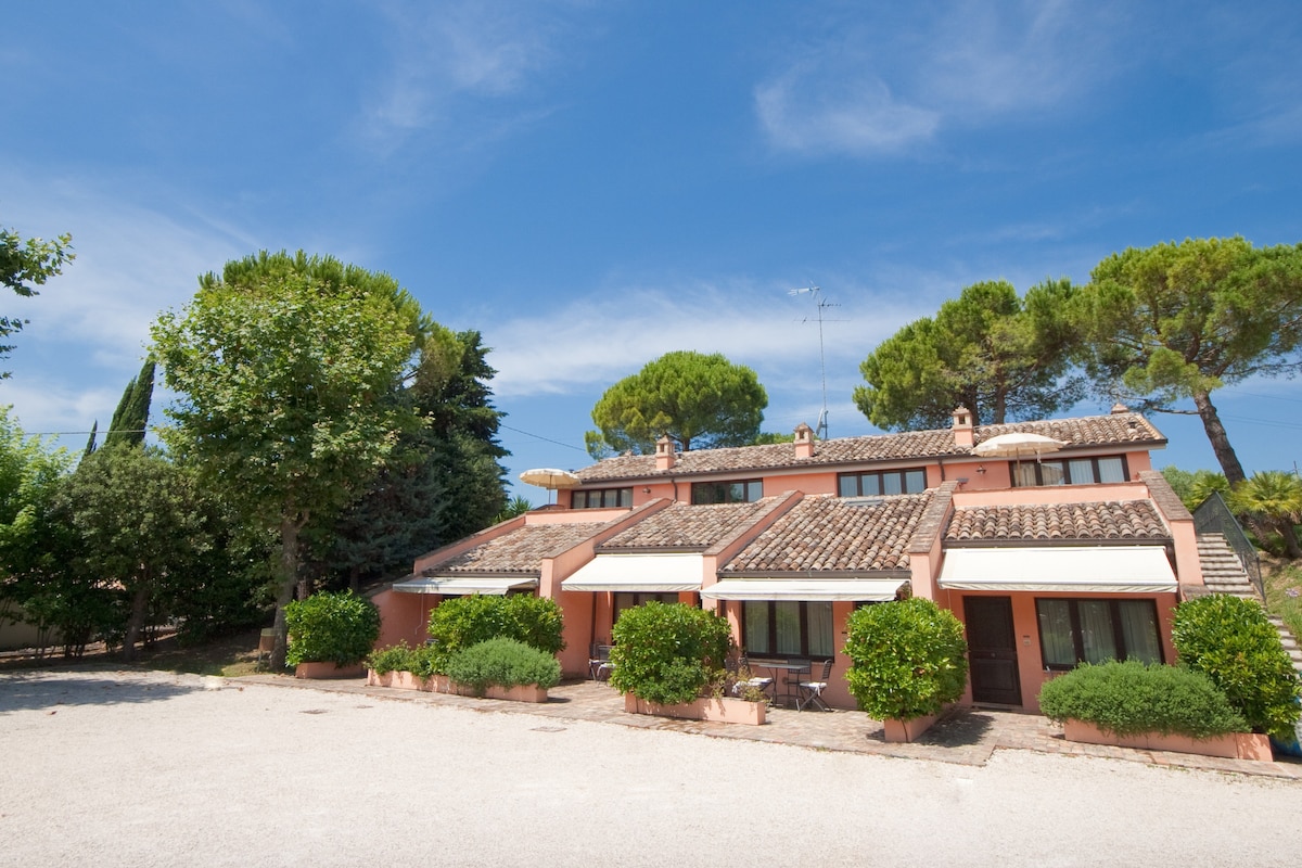 A two-story building features an earthy pink exterior with terracotta roofs. Lush greenery surrounds the entrance, including manicured bushes and tall trees. A gravel driveway leads to the structure, providing ample outdoor space. Clear skies add a bright atmosphere to the setting.
