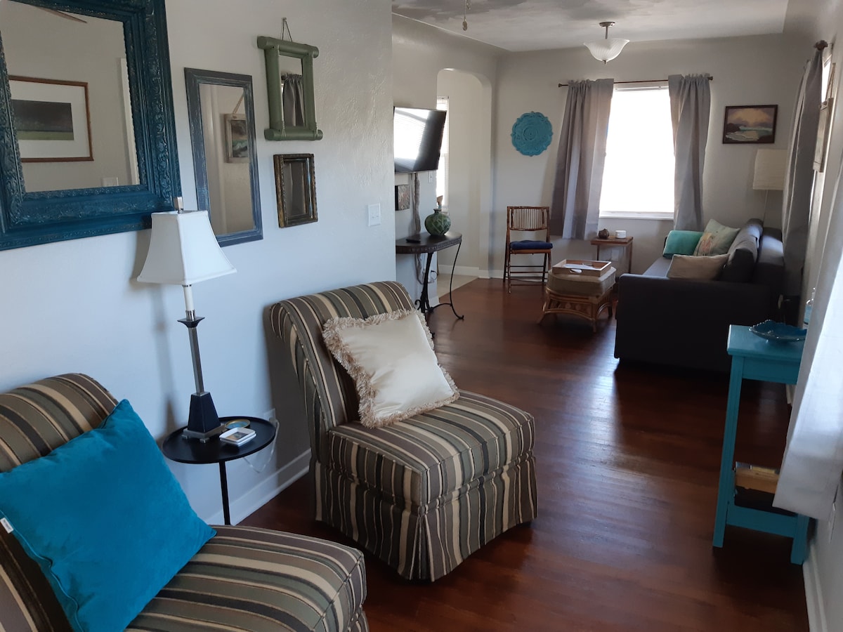 A comfortable living area features striped chairs with decorative pillows and a cozy sofa. Natural light streams through curtains, illuminating the hardwood floor. A side table is positioned beside a lamp, and artwork adorns the walls, enhancing the inviting space.