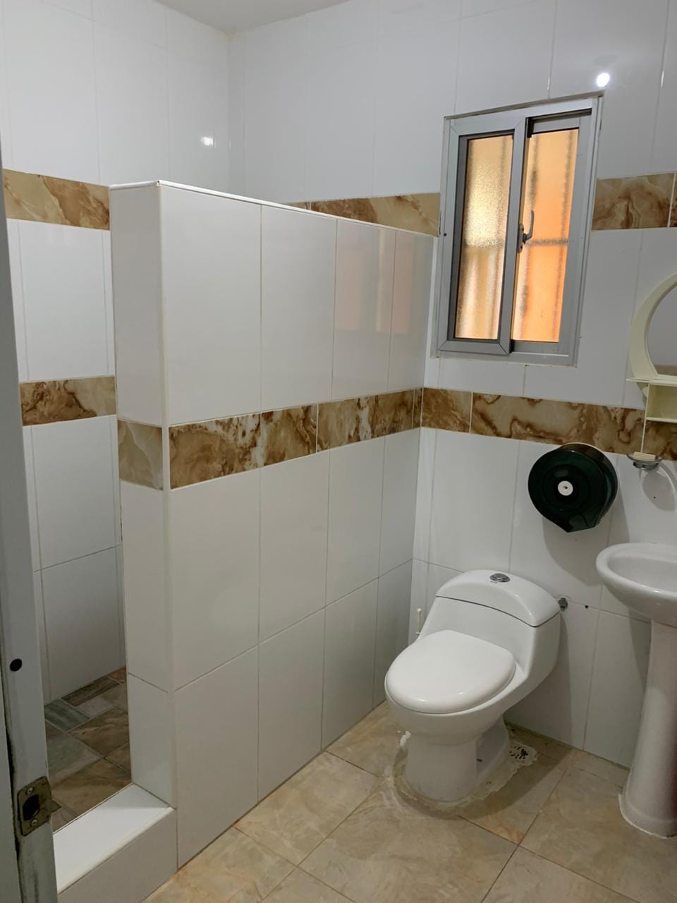 A clean and modern bathroom features white tiled walls with subtle brown accents. A glass shower is located in the corner, and natural light enters through a small window. A pedestal sink and a toilet are positioned for convenience.