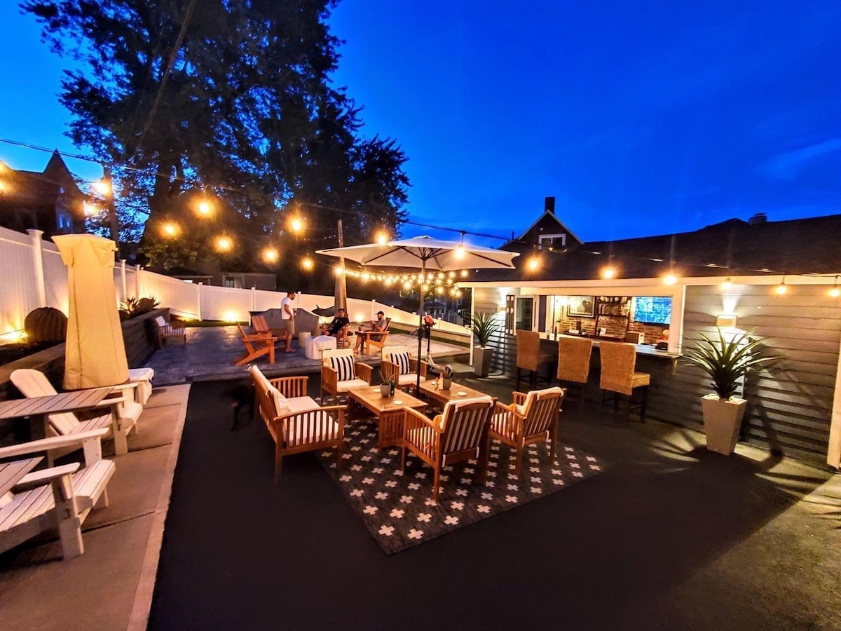 An outdoor patio is illuminated by string lights, showcasing an inviting seating area with multiple chairs and lounge furniture arranged around a central rug. A large umbrella provides shade over a bar area, surrounded by greenery and a bright evening sky.