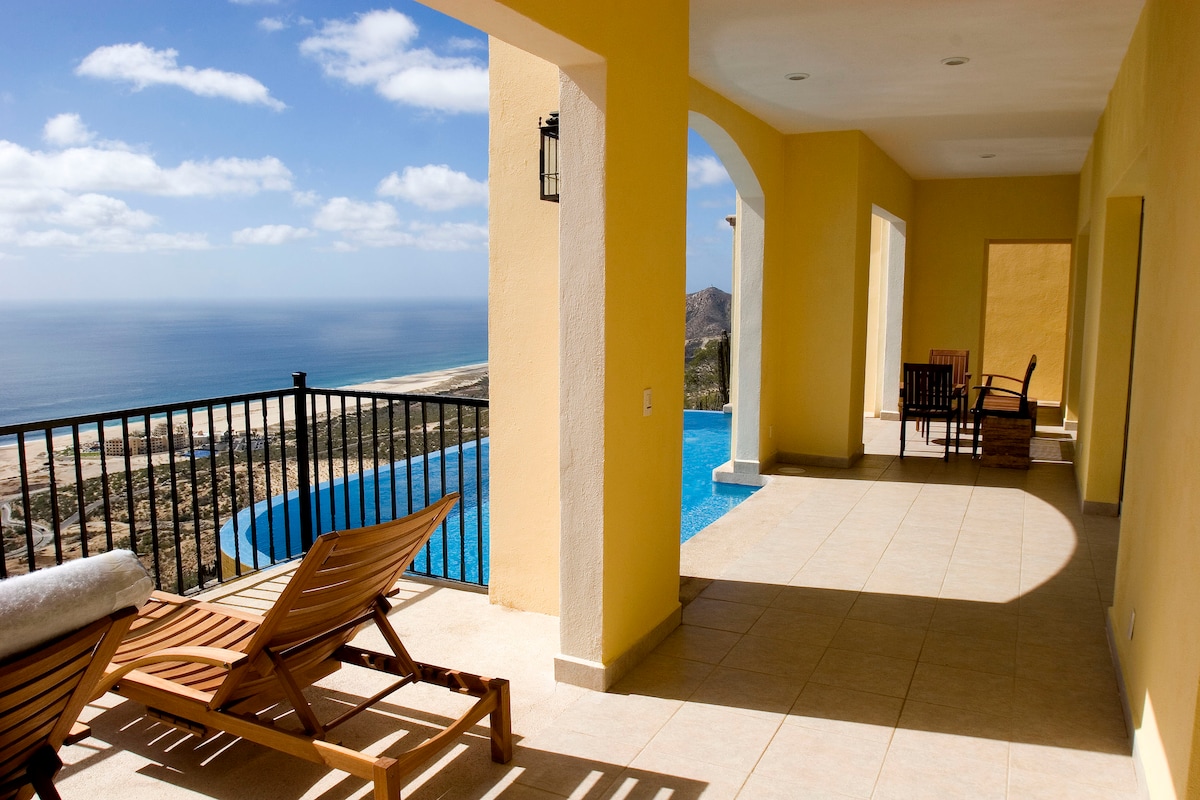 An expansive terrace is displayed, featuring comfortable wooden lounge chairs facing a stunning ocean view. A large infinity pool is positioned nearby, while arched openings lead to an airy interior space. The soft yellow walls enhance the bright atmosphere under a partly cloudy sky.