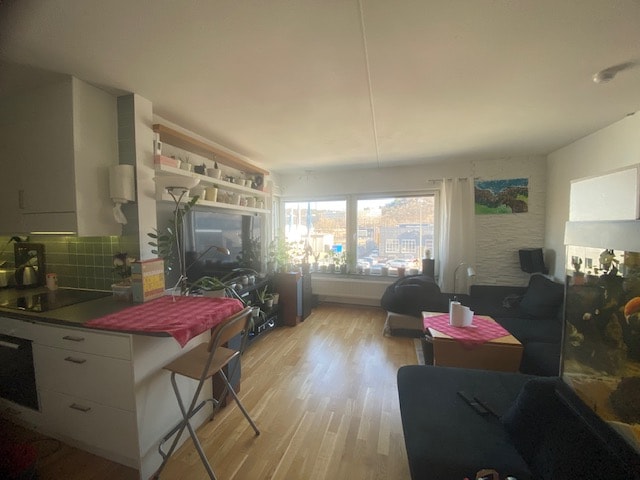 A bright living area features large windows that allow ample natural light. A spacious couch and armchair offer comfortable seating, while a small dining table with two chairs is positioned near the kitchen. Houseplants are placed throughout, adding a touch of greenery.
