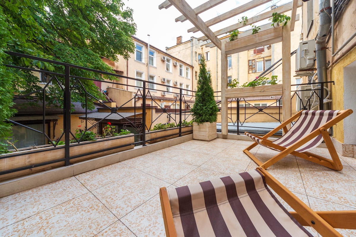 A private terrace features a wooden pergola adorned with greenery, providing shade. Two striped lounge chairs offer a place to relax, overlooking a calm courtyard with buildings in the background. The floor is tiled with a decorative design, enhancing the space's inviting atmosphere.