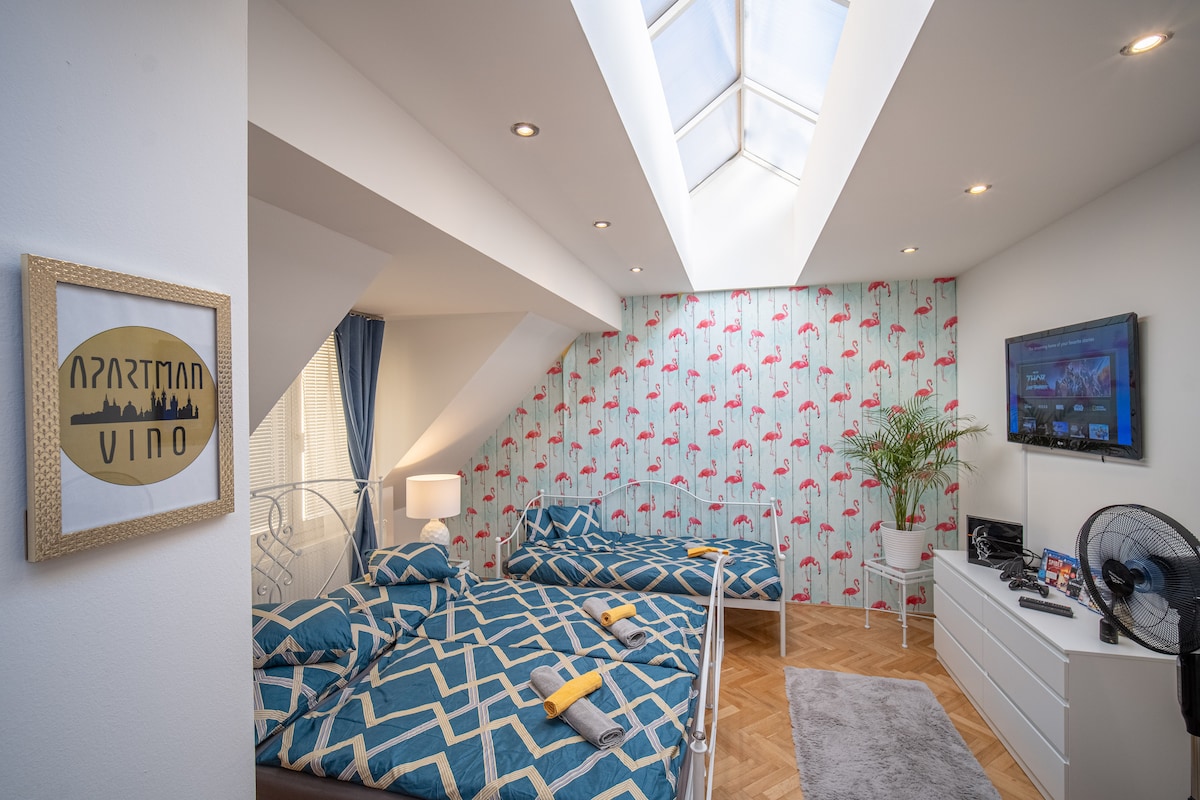 A bright and spacious sleeping area features a skylight allowing natural light to fill the room. Two beds, neatly made with patterned covers, are positioned on a wooden floor. A decorative wall with umbrella motifs adds a playful touch, complemented by a television mounted on a white dresser.