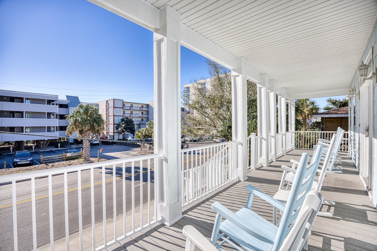 A spacious porch features multiple light blue rocking chairs, inviting relaxation. The area overlooks a road lined with palm trees and nearby buildings. Sunlight filters through the open space, creating a bright, airy environment.