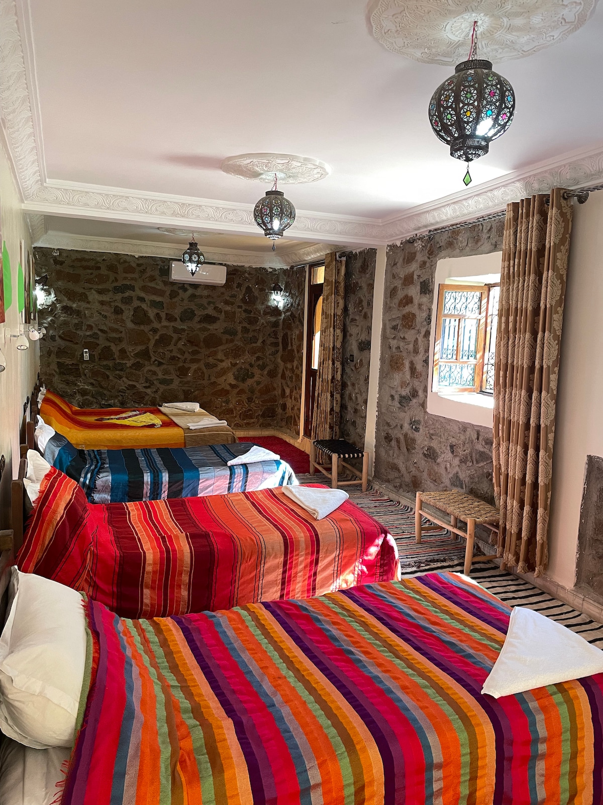 A spacious room is filled with colorful striped bedding on five large beds. The stone walls add a rustic charm, while intricate lanterns hang from the ceiling. Natural light filters through windows, illuminating the warm tones of the decor.