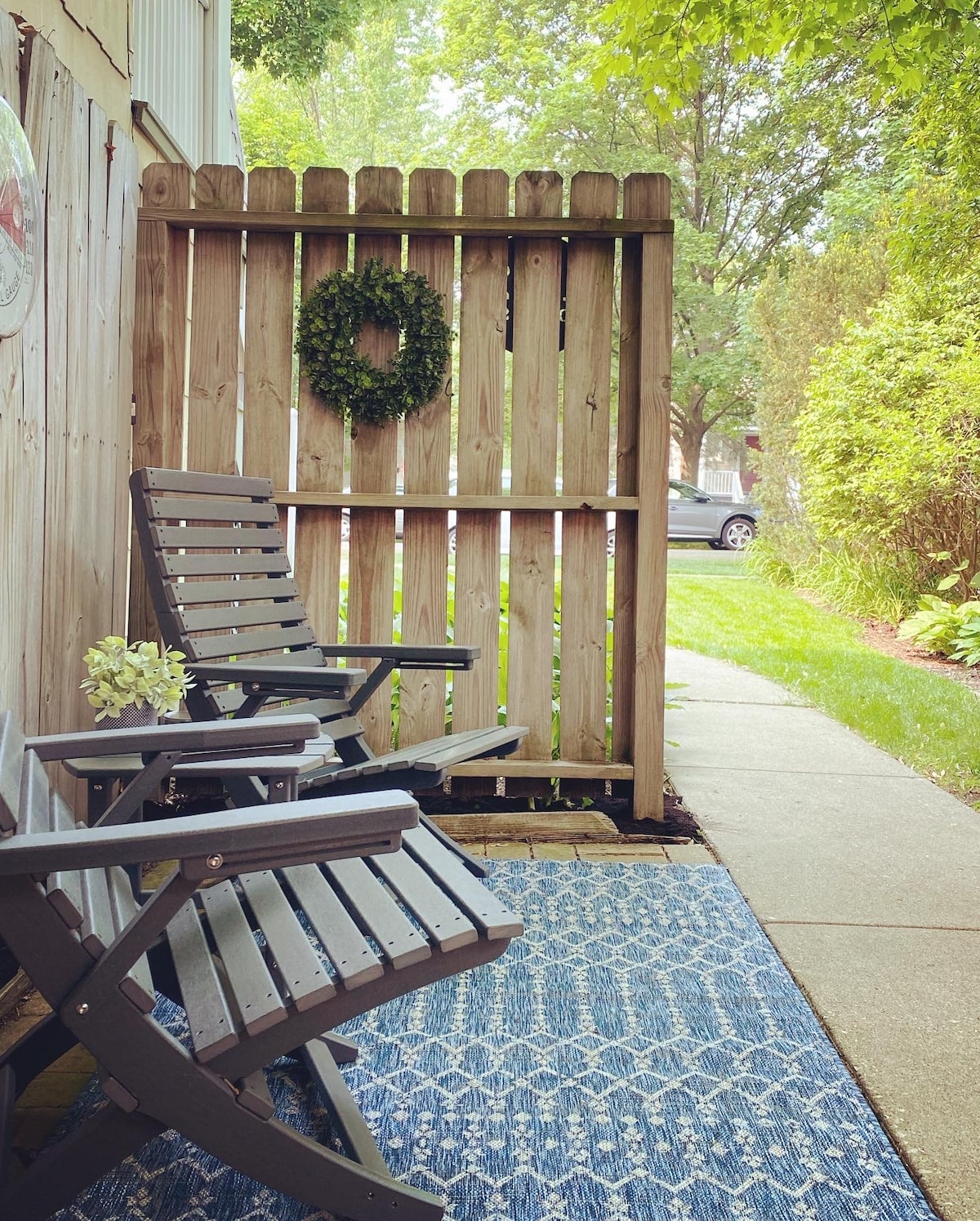 A small outdoor patio area features two wooden chairs positioned on a blue patterned rug. A wooden fence adorned with a circular greenery wreath creates privacy. Lush greenery lines the path, and a concrete walkway leads to the door, inviting relaxation and outdoor enjoyment.