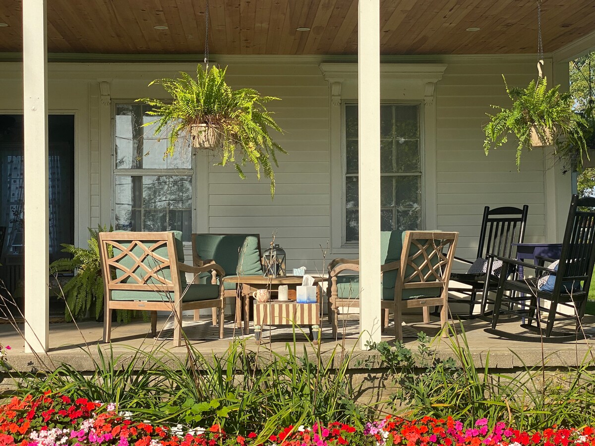 A welcoming front porch includes comfortable seating with soft cushions arranged around a wooden coffee table. Hanging planters with lush green ferns add a touch of nature, while colorful flowers line the base of the porch, enhancing the outdoor space.