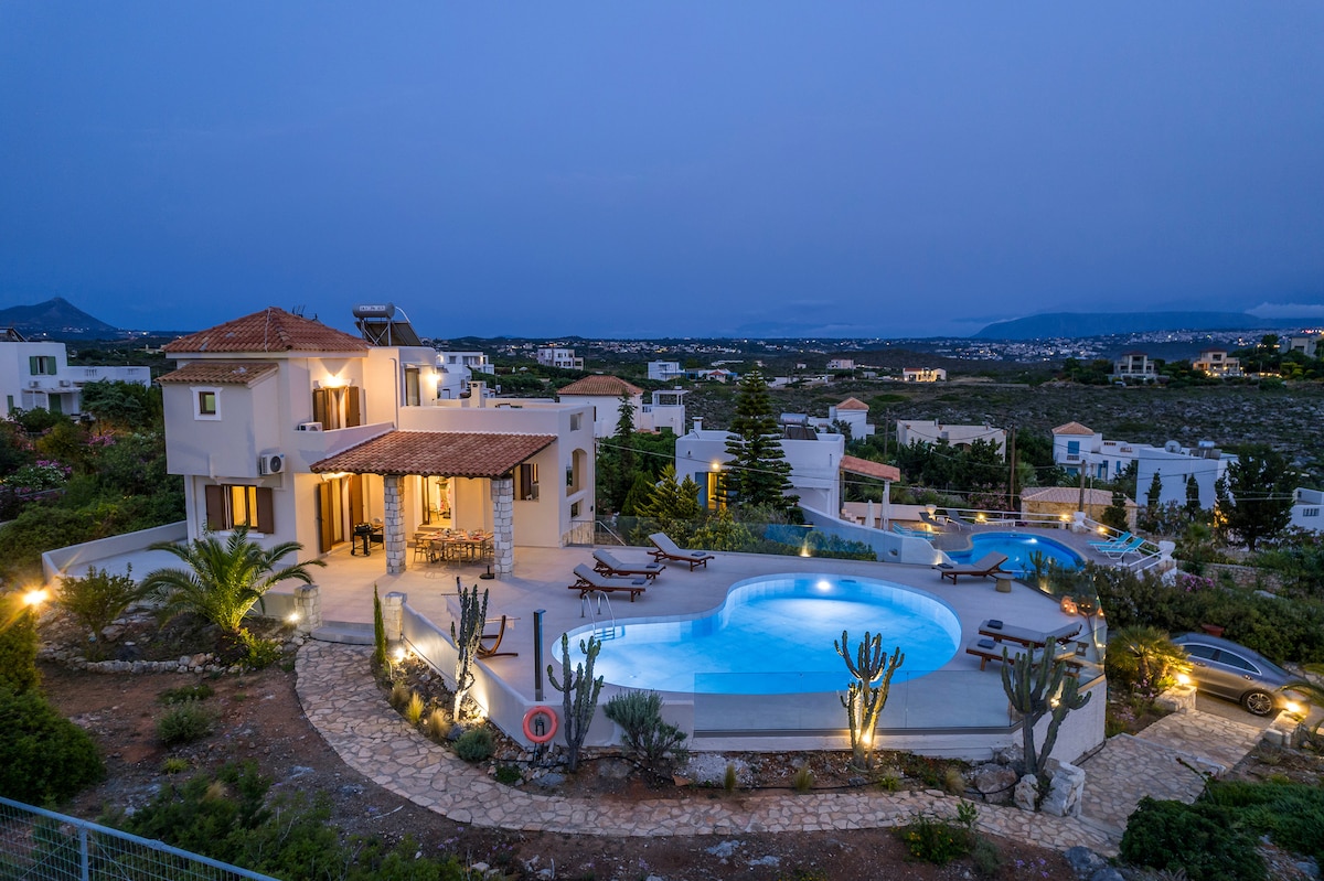 A panoramic view of a luxury villa is presented, featuring a heated pool that glows softly in the evening light. Surrounding greenery and elegant outdoor seating enhance the tranquil setting, with distant views of the village and coastline visible in the background.