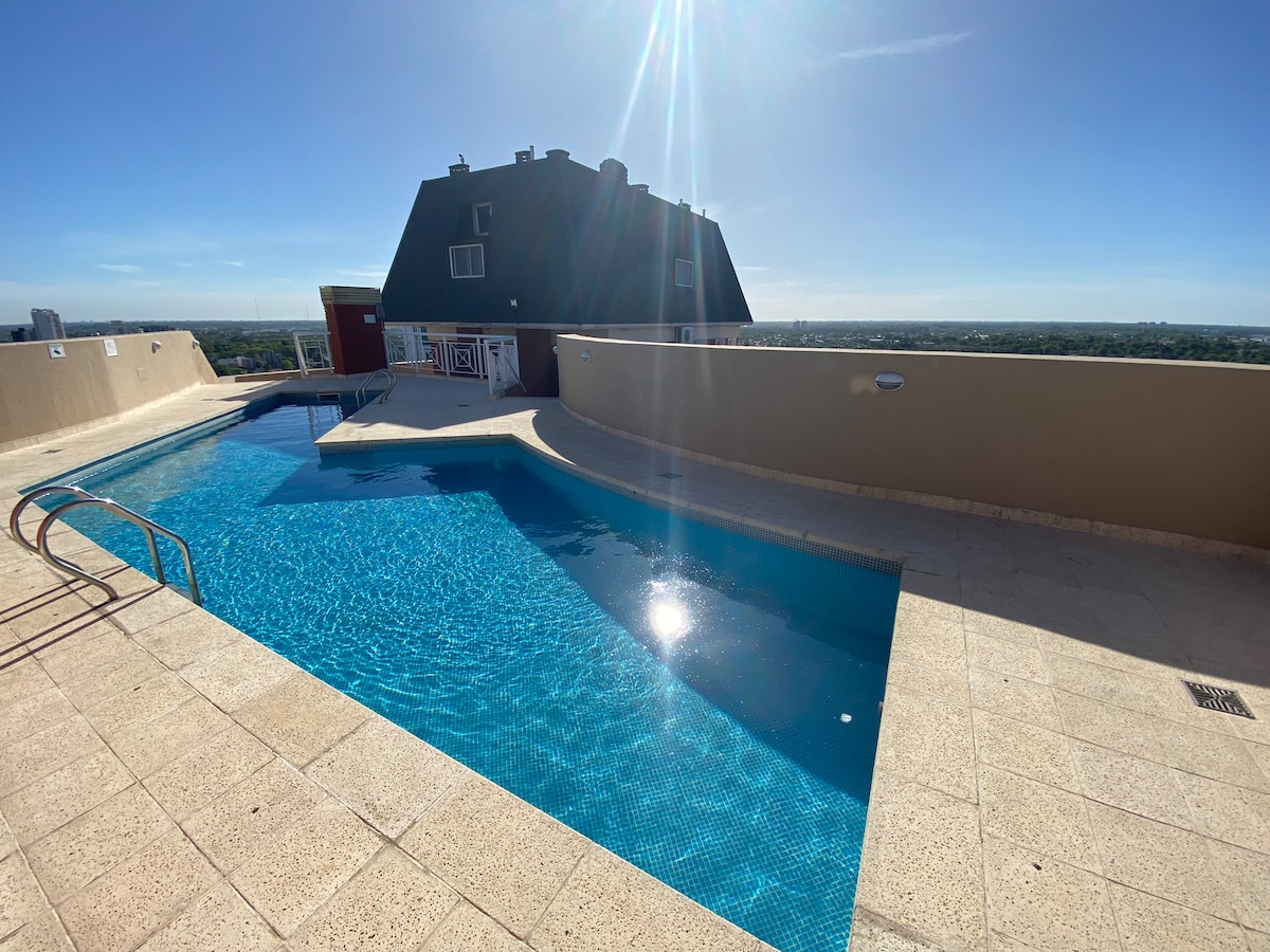 A rooftop pool area is displayed, featuring a large, clear water pool set against a backdrop of a blue sky. The sun reflects off the water's surface, creating a shimmering effect. Surrounding spaces include smooth stone decking and a small staircase leading to the upper patio.