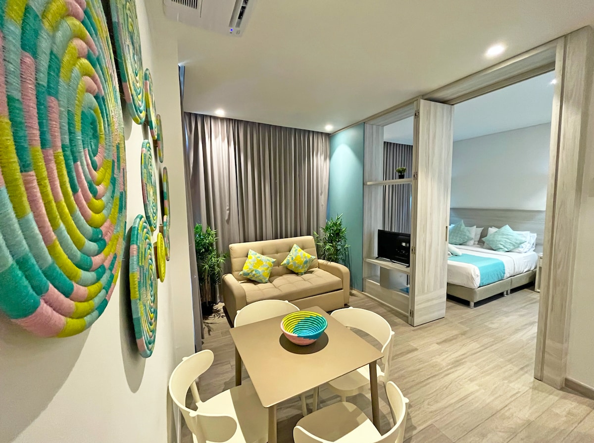 The living area features a light-colored sofa accompanied by a round table and four chairs. Colorful wall decor adds vibrancy to the space. A partially separated sleeping area is visible, showcasing a spacious bed with soft linens and light-colored bedding.