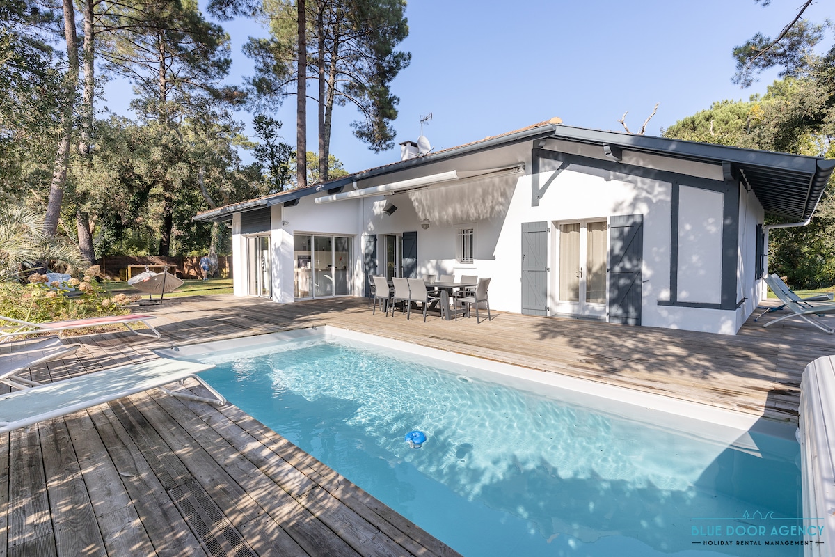 A modern villa with large sliding glass doors leading to a wooden terrace and pool area. Sun loungers are arranged by the serene pool, surrounded by lush greenery. A dining table with chairs is set on the terrace, ideal for outdoor meals.