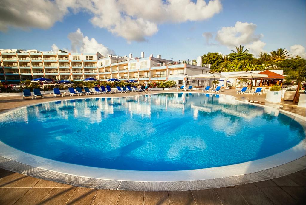 A large circular pool is surrounded by lounge chairs and umbrellas. Reflections of the blue sky and clouds can be seen on the water's surface. Nearby buildings and greenery frame the pool area, creating a serene and inviting environment.