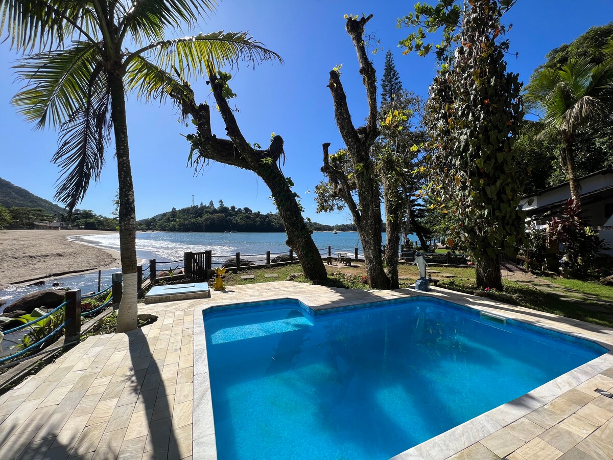 A crystal-clear swimming pool is framed by lush greenery and swaying palm trees, with a sandy beach and tranquil water visible in the background. The pool area features a tiled deck, providing an inviting space for relaxation and enjoyment.