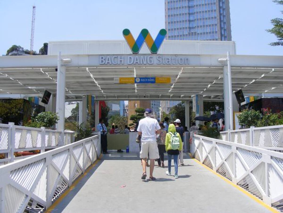 The entrance of Bach Dang Station is displayed, featuring a white structure with a distinctive triangular roof design. Colorful signage indicates the station's name, and a wide walkway leads towards the entry. Visitors can be seen approaching the station amidst a sunny day.