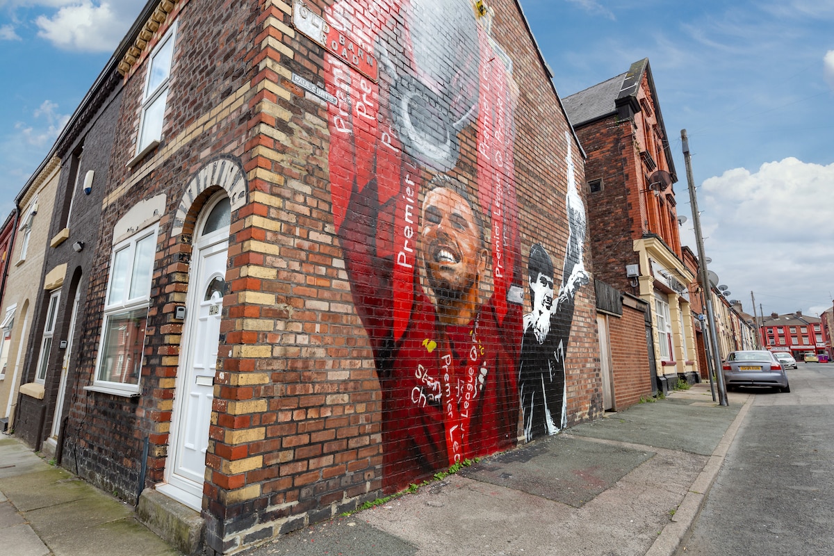 An exterior wall of a brick house features a vibrant mural created by local artist Paul Curtis, showcasing a Liverpool football player with striking red and black hues. The surrounding street displays a mix of residential buildings, contributing to the local atmosphere.