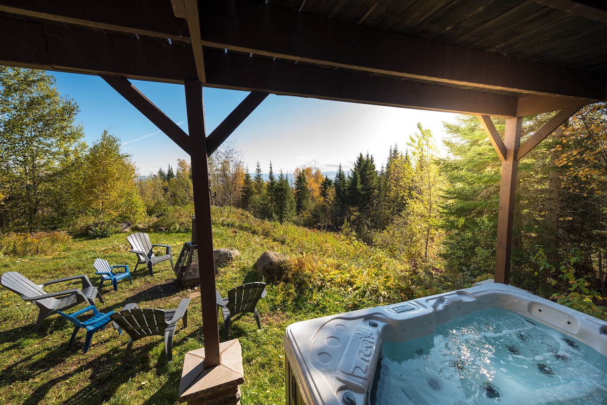 A hot tub is positioned under a wooden overhang, offering views of the surrounding greenery and distant trees. Several Adirondack chairs are arranged within the outdoor space, inviting relaxation amidst the natural setting.