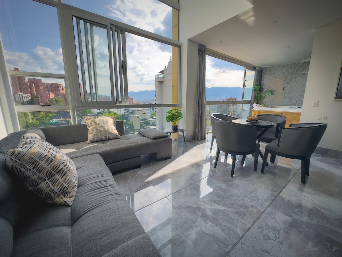 A modern living area features a comfortable gray sectional sofa, accented by a patterned pillow. Floor-to-ceiling windows provide a panoramic view of the city, allowing natural light to fill the space. A dining table with black chairs is positioned nearby on polished tile flooring.