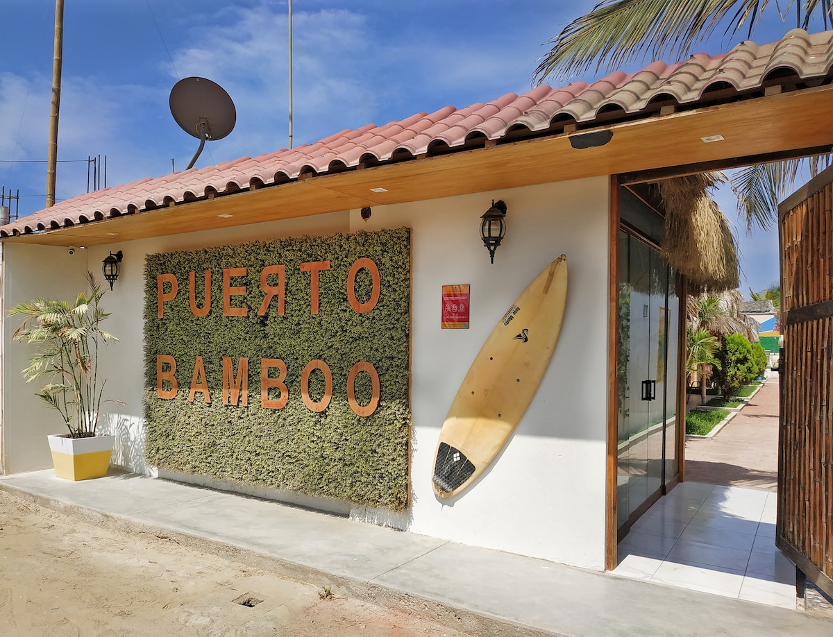 The exterior of Puerto Bamboo features a welcoming entrance adorned with lush greenery and a prominent sign displaying the name. A surfboard is mounted on the wall beside the door. The pathway leads toward the inviting interiors of the property, framed by palm trees.