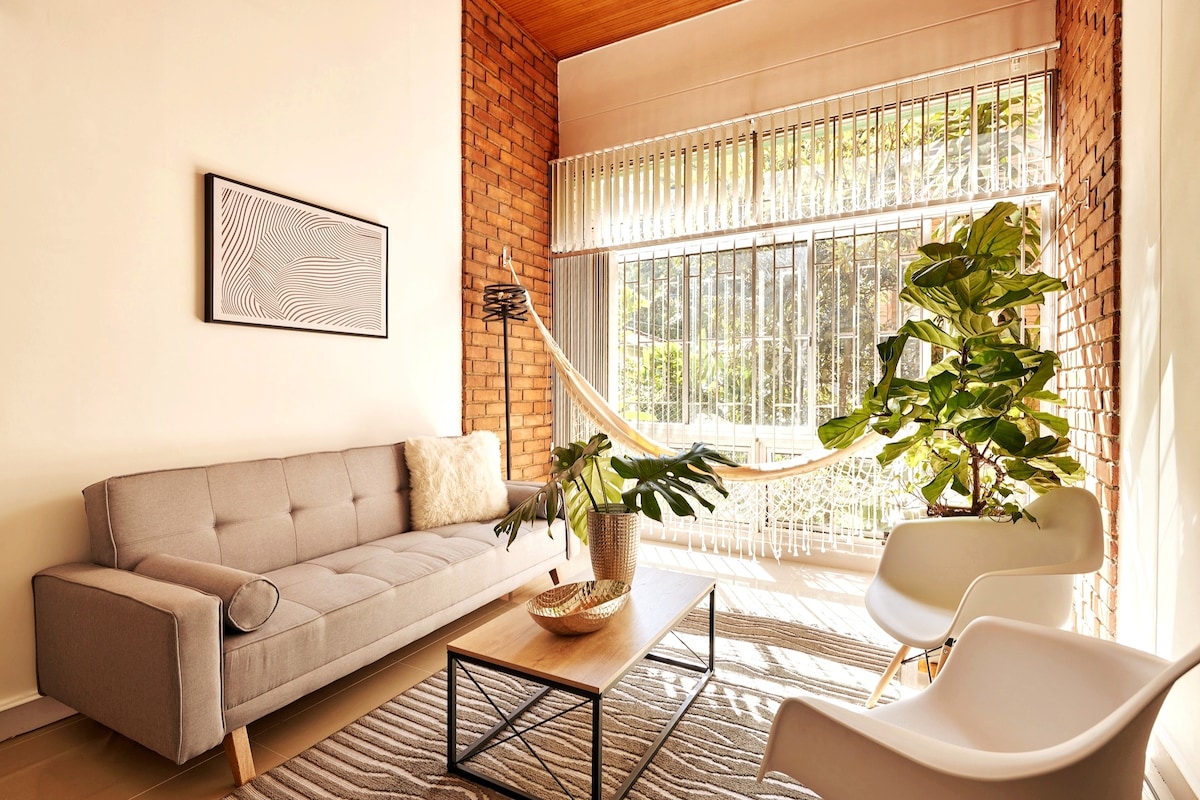A light-filled living room showcases a modern gray sofa and a pair of comfortable white chairs. A stylish coffee table sits in the center, complemented by decorative elements. Lush greenery can be seen through the large window, which is framed by elegant window coverings.