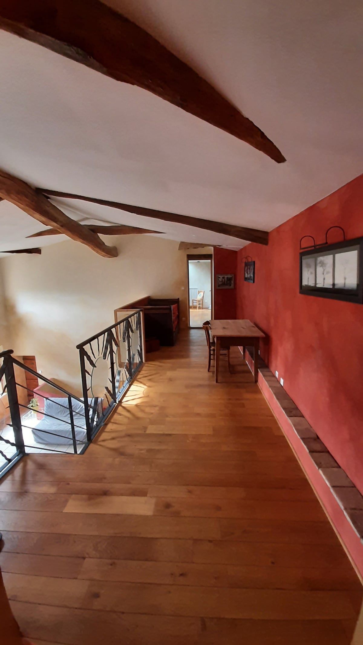 A hallway is shown reflecting warm tones from the wooden flooring and walls. Exposed beams add character to the ceiling. A railing provides safety, with a wooden table positioned against the wall. Art pieces and photographs are displayed, contributing to the space's charm.