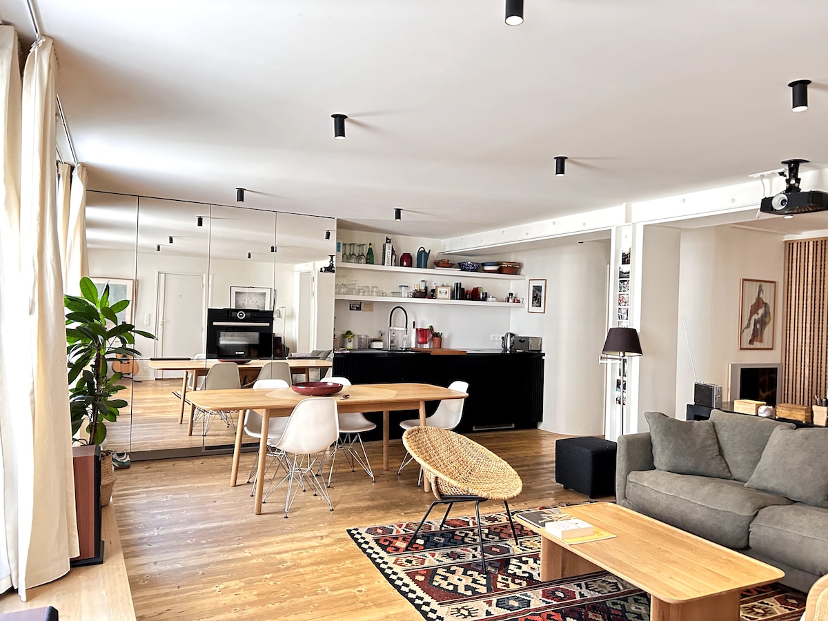 A spacious living area features a large dining table surrounded by chairs, set against a modern kitchen backdrop with open shelving. A comfortable sofa and coffee table invite relaxation, while a patterned area rug adds warmth. Natural light filters through soft curtains.