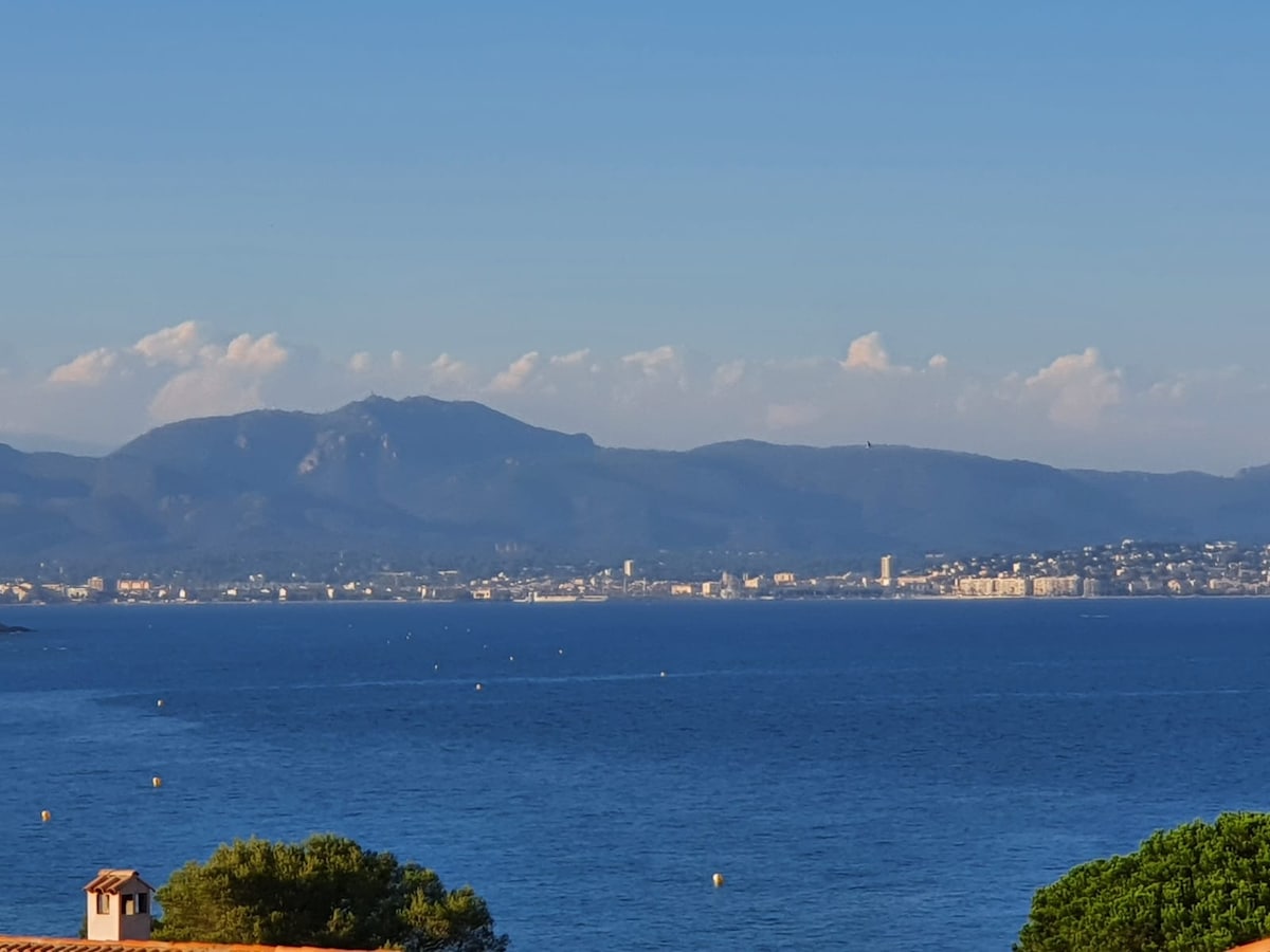 A panoramic view of the coastline is presented, showcasing the tranquil blue waters seamlessly meeting the distant shoreline. Lush greenery is visible in the foreground, while gentle mountains rise in the background under a clear blue sky dotted with soft clouds.