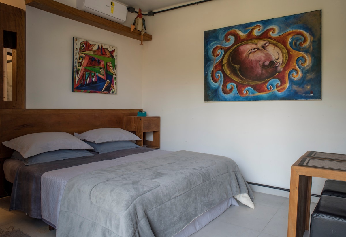 A cozy sleeping area features a spacious bed dressed in gray bedding, complemented by two textured pillows. Artwork adorns the walls, displaying vibrant colors. A simple wooden nightstand and table provide functional decor, enhancing the room's modern atmosphere.