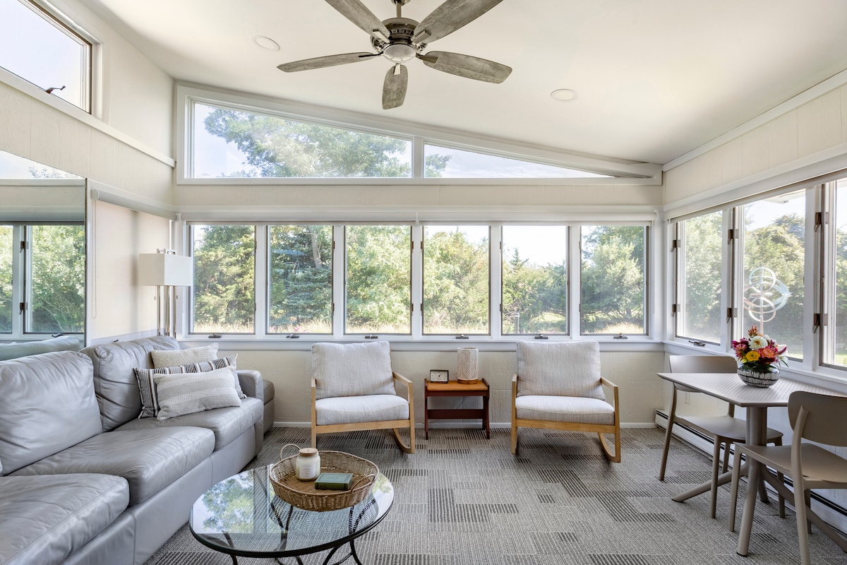 A sunlit room features large windows with a view of surrounding greenery. Comfortable seating includes a plush sofa and two armchairs, with a small table at the center. A ceiling fan hangs above, enhancing the spacious feel of this inviting space.