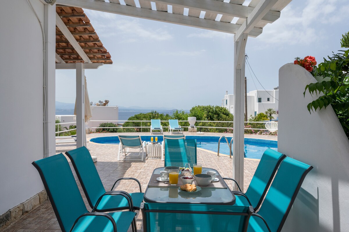 A shaded terrace area features a glass dining table surrounded by four teal chairs. Breakfast items, including pastries and drinks, are displayed on the table. In the background, a spacious swimming pool and surrounding greenery can be seen, with distant sea views under a clear sky.