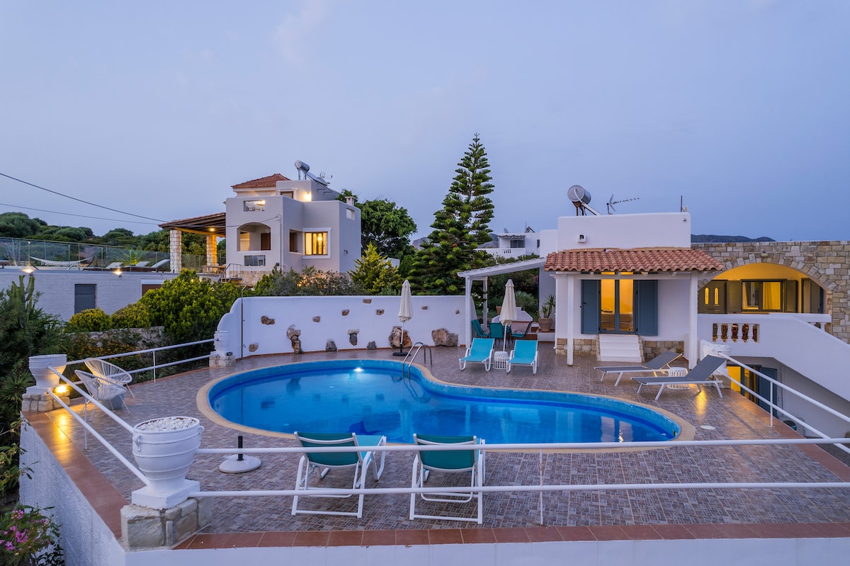 The outdoor pool area features a spacious crystal-clear pool surrounded by sunbeds and umbrellas. A covered terrace area provides shade, and the villa is framed by lush greenery and surrounding buildings, creating a serene environment for relaxation.