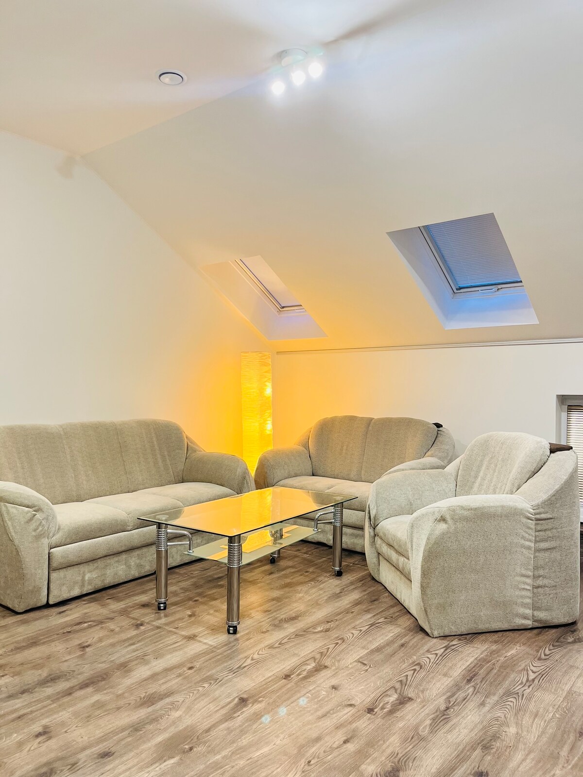 A softly lit seating area is depicted, featuring two comfortable sofas and an armchair arranged around a glass-topped coffee table. Natural light filters through two skylights, creating a warm ambiance against the light-colored walls.