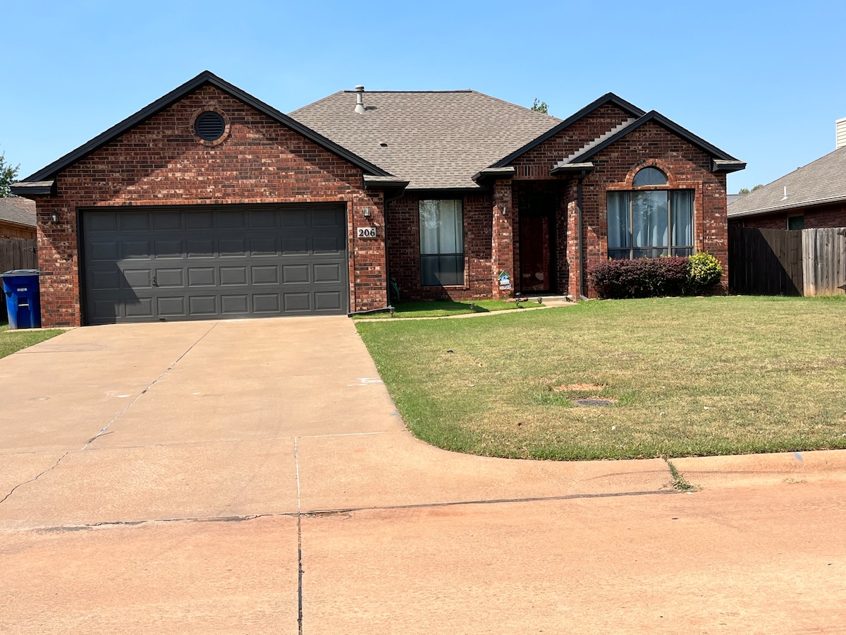 The exterior of a single-story brick home is shown, featuring a neatly manicured front lawn and a paved driveway. A two-car garage and large windows contribute to the welcoming facade. A privacy fence can be seen in the background.