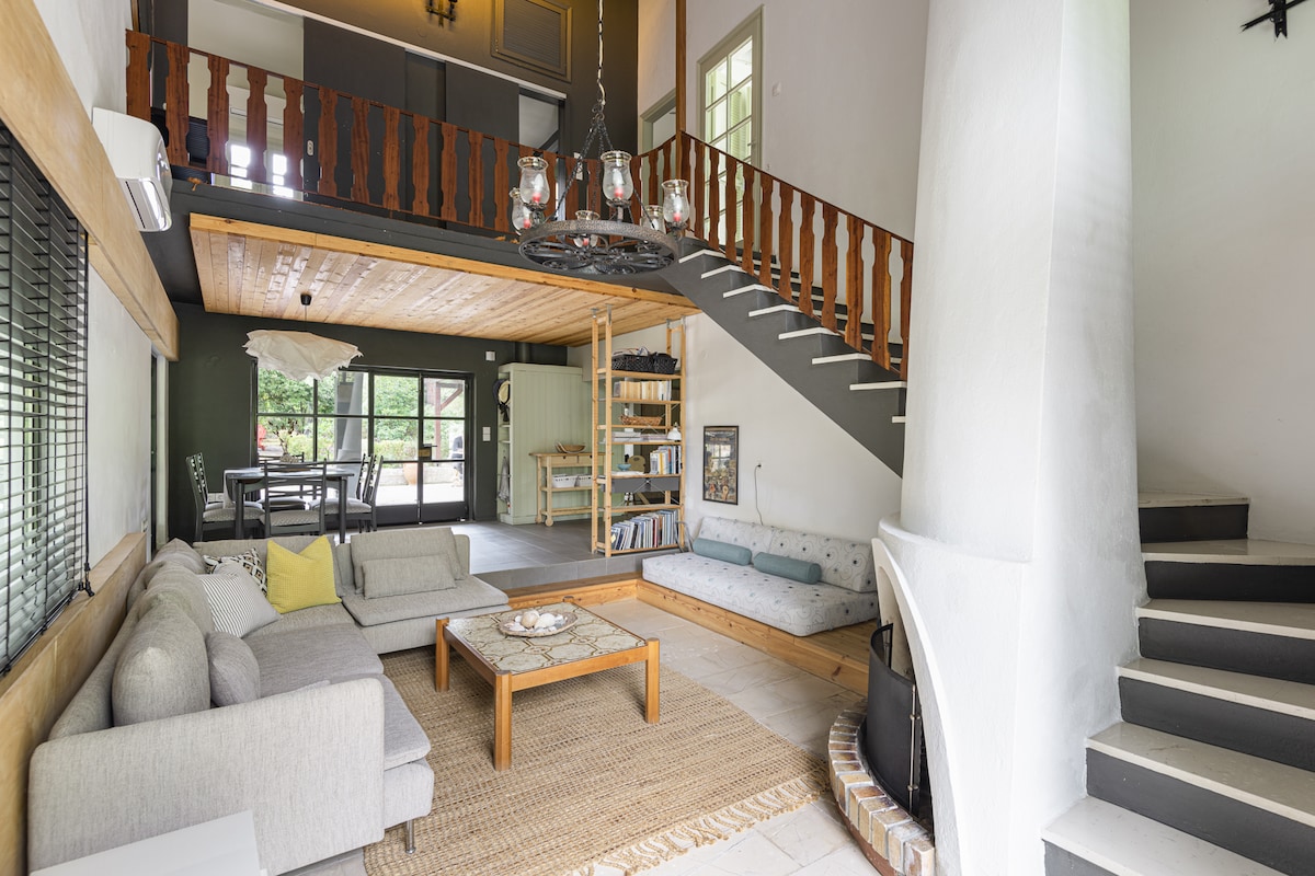 The living area features a spacious layout with a cozy seating arrangement, including neutral-toned sofas. A wooden coffee table sits centrally, surrounded by a woven rug. A staircase leads to the upper floor, and large windows provide natural light, enhancing the welcoming atmosphere.