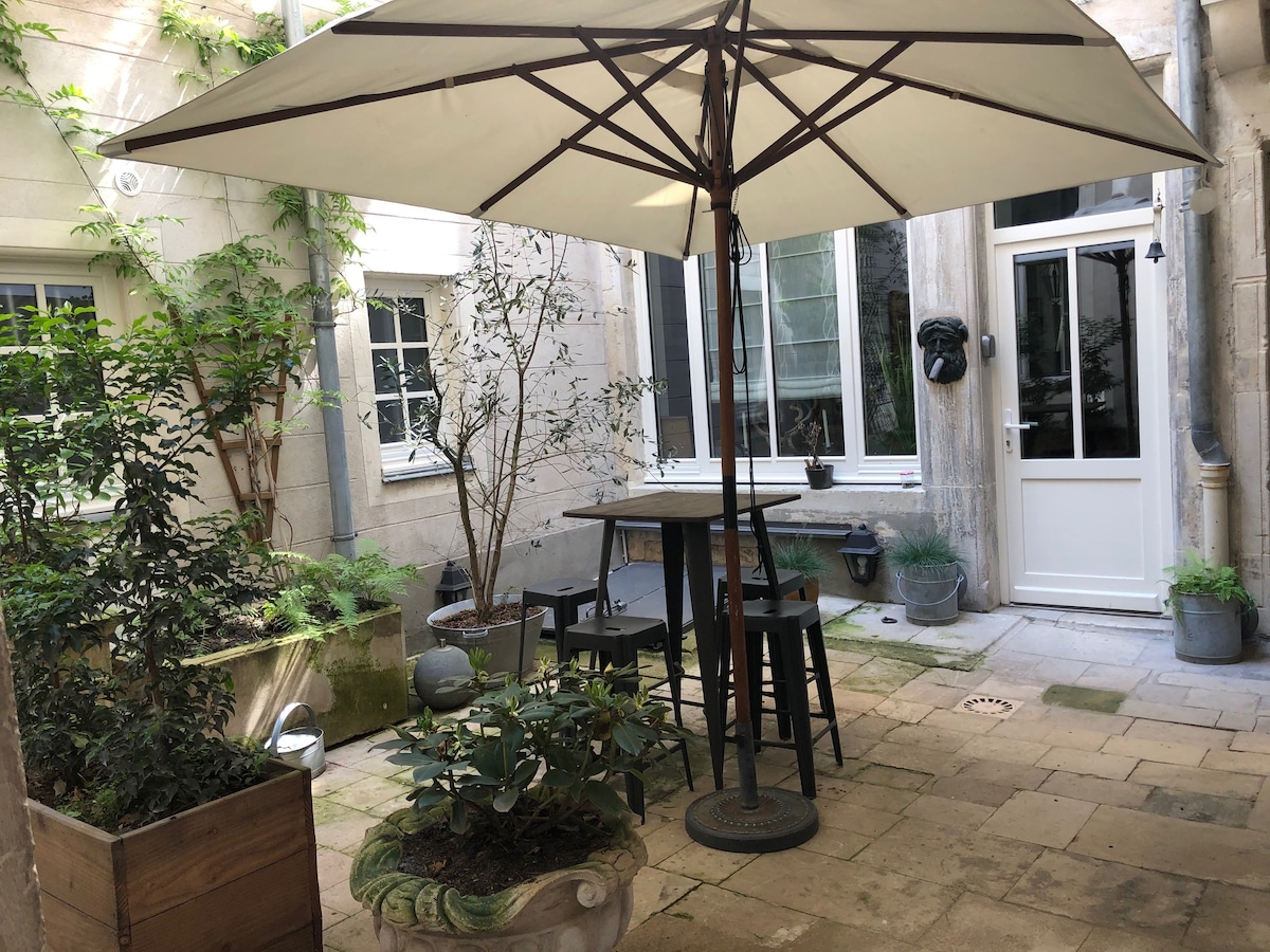 A private courtyard is featured, surrounded by stone walls and greenery. A large umbrella provides shade over a tall table and stools. Planters with plants are positioned around the space, enhancing the outdoor atmosphere.