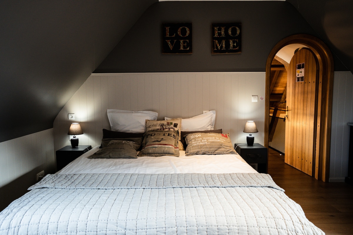 A spacious bed is adorned with decorative pillows, positioned between two bedside tables featuring soft lighting. The room is decorated with muted tones, complemented by wooden elements in the archway leading to another area.