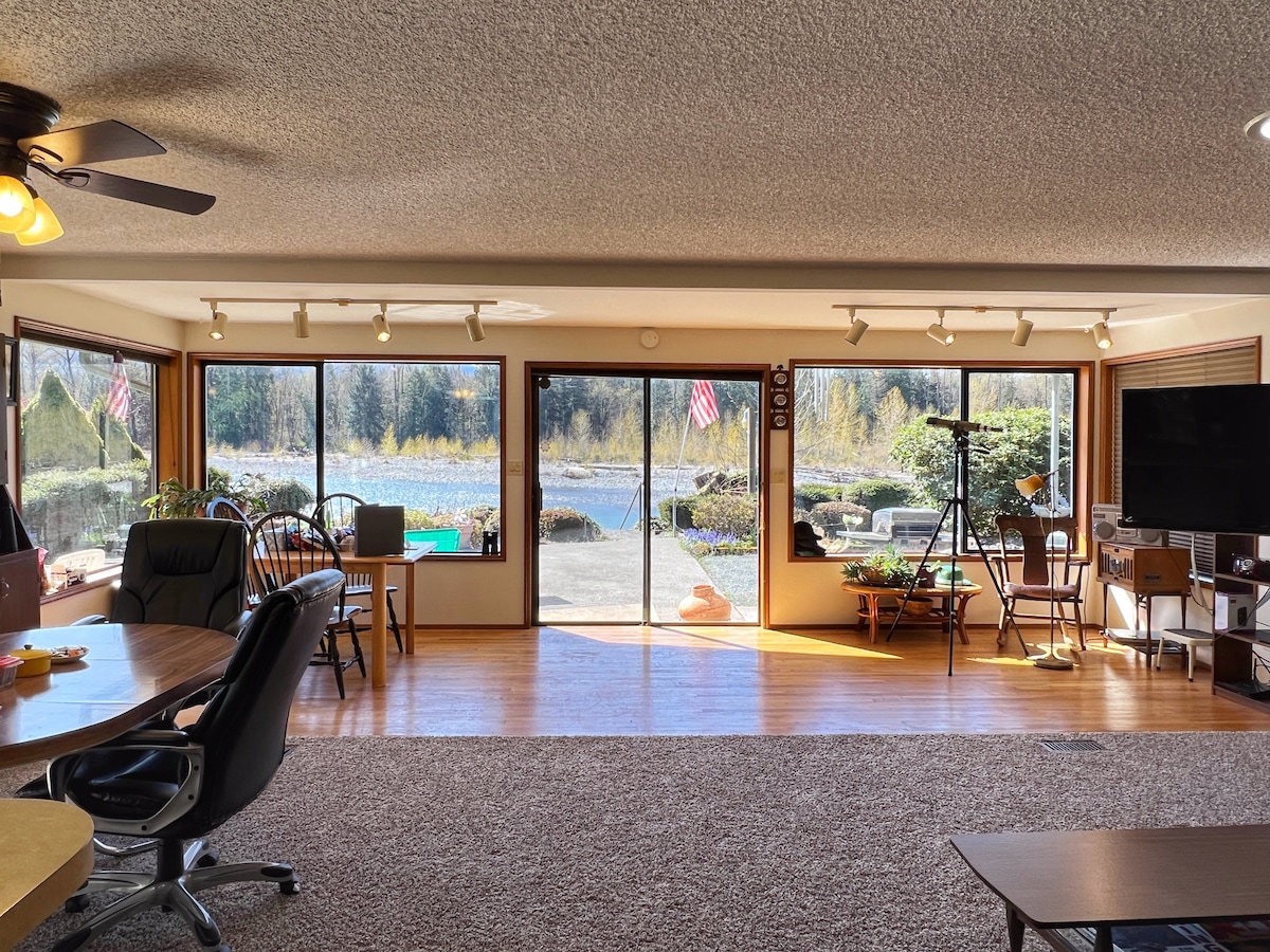 Large picture windows provide expansive views of the waterfront and surrounding nature. The open living area features two dining spaces, a cozy seating arrangement, and a telescope positioned for birdwatching. Natural light illuminates the room, highlighting its wood accents and inviting layout.