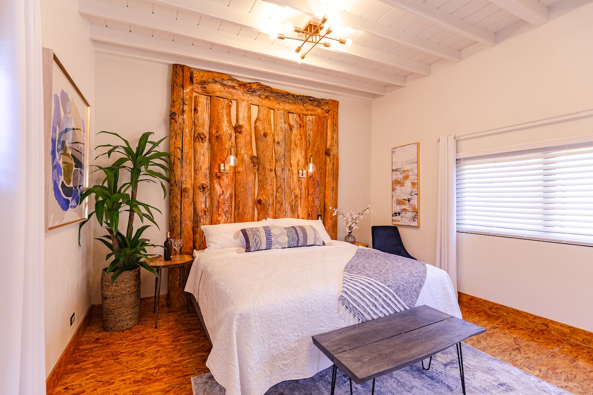A spacious bedroom features a large bed with a textured white coverlet, set against a striking wooden accent wall. Soft lighting complements the room's calming palette, while a small table and a chair add functionality. A plant in the corner enhances the natural feel.