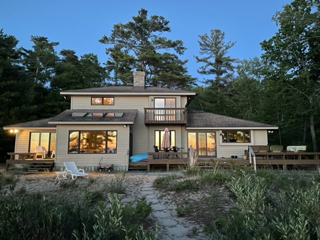 A two-story house is nestled among trees, featuring multiple large windows reflecting the twilight sky. A spacious deck is accessible from the main level, furnished with outdoor seating. Soft lighting from the interior casts a warm glow, enhancing the inviting atmosphere of the property.