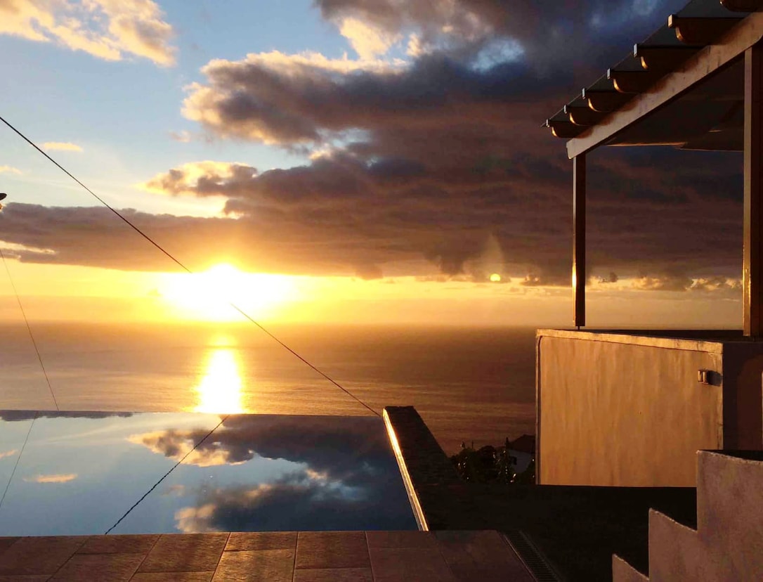 The infinity pool reflects the vibrant sunset, creating a seamless connection between water and sky. A shaded outdoor area with a wooden pergola stands alongside, enhancing the coastal view. The horizon is filled with warm hues of orange and gold, complemented by distant clouds.