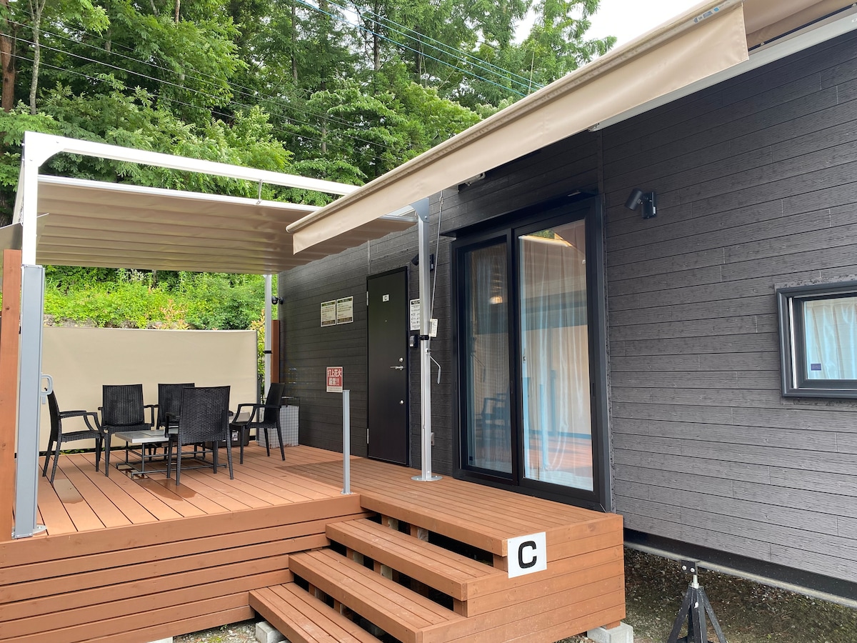 An outdoor seating area is presented on a wooden deck, featuring a table surrounded by four chairs. An awning provides shade, while a door leads into the trailer house. Lush greenery is visible in the background, enhancing the tranquil environment.